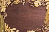 Dark smooth wooden background surrounded by dry pasta in various types and lengths. Includes copy space.
