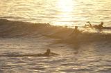 A person riding a surf life saving board rides a wave past other surfers in bright, golden sunset light.