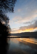 Sunset over the placid water of Lake Windermere in the English Lake District, Cumbria, England with trees in the foreground silhouetted against a colourful sky reflected in the lake below