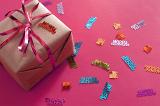 Over View of Gift Wrapped in Brown Paper and Pink Foil Ribbon Surrounded by Birthday Themed Confetti on Hot Pink Background