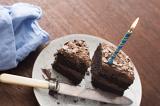 Chocolate cake with lit blue candle on white plate next to dull knife and cyan cloth napkin atop wooden table