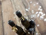 Angled Over Head View of Two Mini Champagne Bottles and Glasses Surrounded by Silver Snowflake Shaped Confetti on Wooden Background
