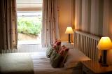 View from the side of a comfortable double bed overlooking a window with a view of greenery with two lamps burning on bedside tables