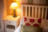 Bedside table and lamp alongside a bed with a simple slatted wooden headboard with floral cushions on the counterpane