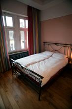Metal frame double bed in an austere bedroom interior with a bare wooden floor and high windows overlooking an urban building