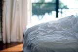 Empty messy unmade bed with white bed linen and crumpled sheets, close up view in front of a bright window