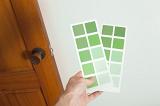 Man holding up cards of green paint swatches against a white wall in his house as he makes a decision on colors to redecorate the interior