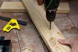 Carpenter drilling a hole in a plank of wood using a handheld electric power drill as he works on a brick floor