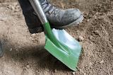 Man wearing a boot digging with a green metal garden shovel in the soil pushing down with his foot