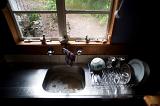 View of a stainles steel kitchen sink with dirty water and washing up on the side in the shadows in front of a window overlooking a back garden