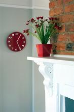 Closeup of a white painted fireplace with a pot of red flowers on the mantelpiece and an ornamental red clock mounted on the wall alongside in an interior decor concept