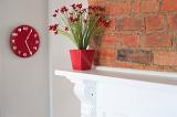 Decorative red flowers in a rustic lounge standing on a cupboard against a face brick wall with a stylish red clock on the wall alongside