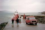 Car disembarking from a ferry onto dry land on a wet rainy day at the coast with people standing around watching