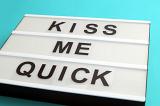 Kiss Me Quick sign with black letters on white box, viewed in close-up and placed on cyan background