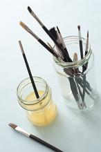 Top down view of various sized artist paintbrushes in jars and thinner solution over white table