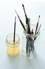 One dirty paint brush in cleaning solution next to jar full of various sized tips over white background with shadow