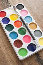 New box of watercolor paints and paintbrush lying open on a wooden desk displaying the range of colors in round pots, high angle view